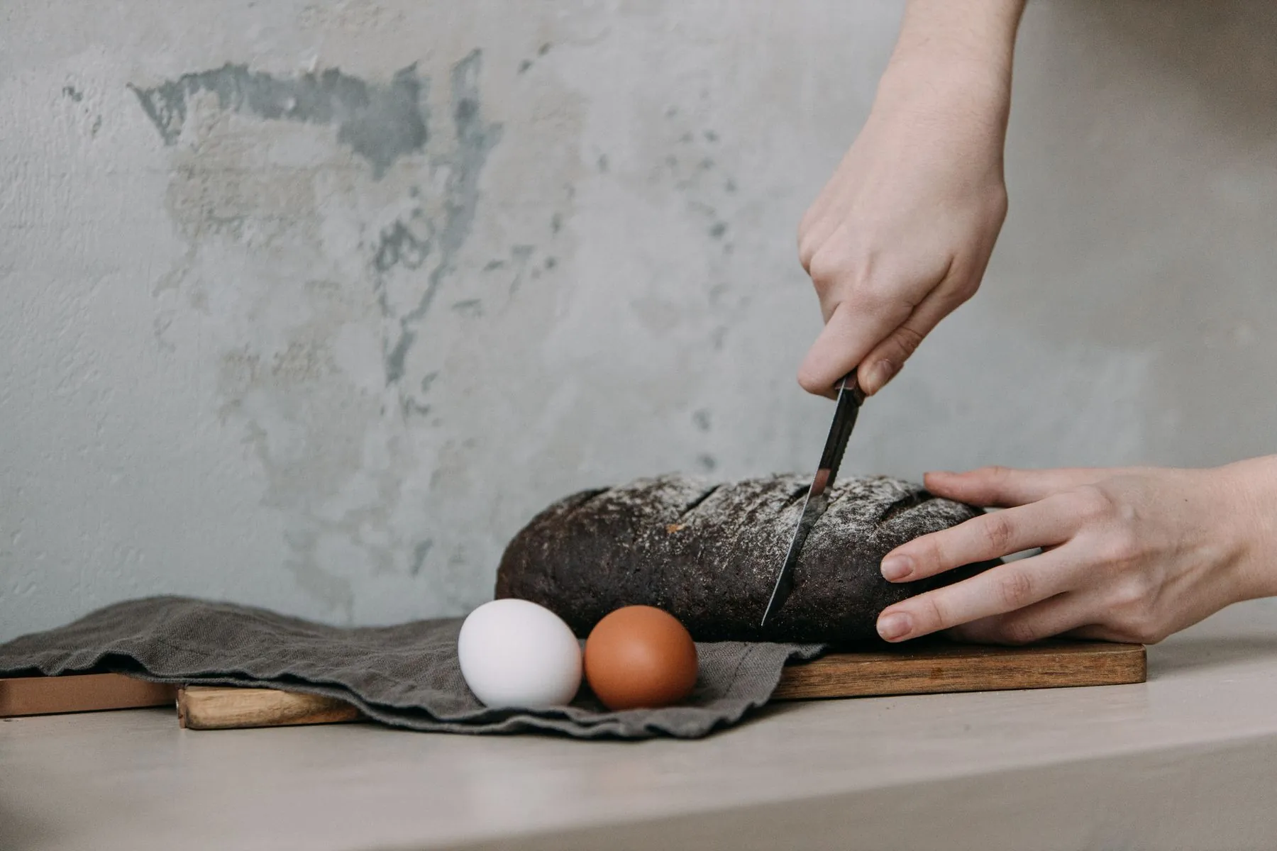 Hands slicing a dark crusted loaf on a linen cloth beside two eggs